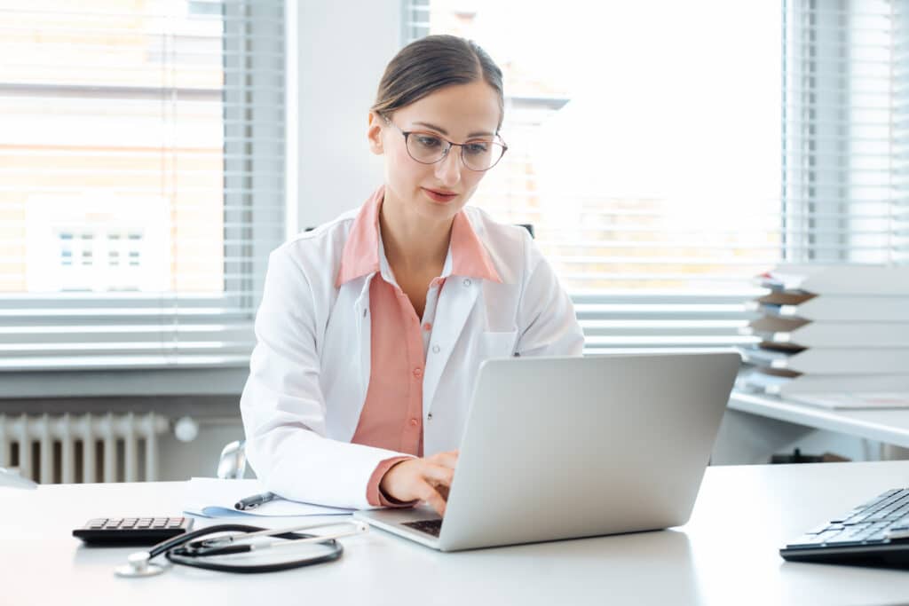 Doctor,Doing,Paperwork,On,Computer,Sitting,On,Desk,In,Her